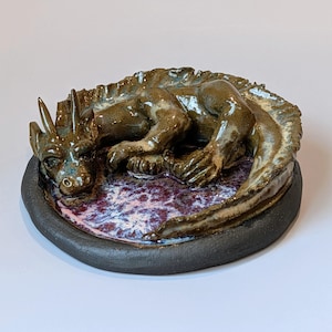 May include: A ceramic sculpture of a dragon resting on a circular base. The dragon has a glossy brown finish with textured scales and horns. It lies curled atop a flat, circular surface featuring a mottled purple, white, and blue glaze.