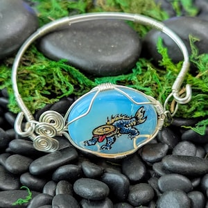 May include: A silver wire bracelet with a blue oval centerpiece. The centerpiece features a blue and red creature design. The bracelet is set against a backdrop of black stones and green moss.