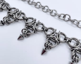Könnte beinhalten: Eine silberne Kettenkette mit einer Reihe von dreieckigen Anhängern. Jeder Anhänger hat eine spitze Spitze und besteht aus ineinandergreifenden Metallringen.