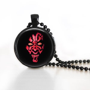 May include: Black pendant necklace featuring a round pendant with a red and black graphic of a character's face. The character has red markings and yellow eyes. The necklace has a black chain.