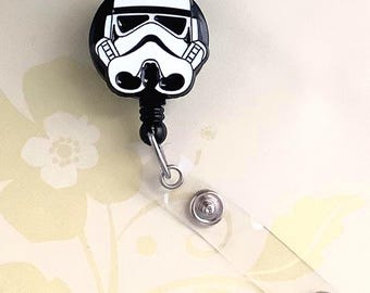May include: A black and white Stormtrooper badge reel. The reel features a detailed image of a Stormtrooper helmet. The reel has a black cord and a clear plastic strap with two snap closures.