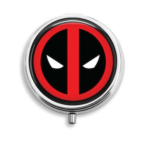 May include: A round, silver-colored pill box with a red and black Deadpool logo on the lid. The logo features a red circle with a black outline, a black mask with a red line down the middle, and two white eyes.