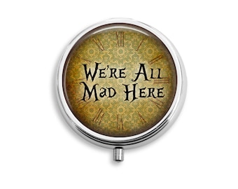 May include: A silver round compact with a green and gold patterned design. The compact has the text "We're All Mad Here" in black.