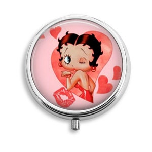 May include: A round, silver compact mirror with a pink background and a cartoon image of a woman blowing a kiss. The image is surrounded by red hearts.