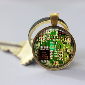 May include: A bronze key chain with a round glass charm featuring a green circuit board with gold-colored components. The circuit board has text including "SHARP JAPAN" and "IR3P90Y1".
