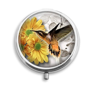 May include: A round silver compact with a hinged lid. The lid features a colorful image of a hummingbird in flight, hovering near yellow flowers with white and gray accents.