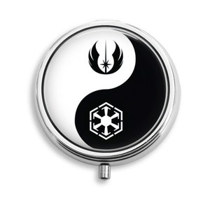 May include: A round, silver pill case with a yin-yang design. One side is white with a black symbol, the other is black with a white symbol. The symbols are from a popular science fiction franchise.