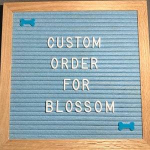May include: A blue felt letter board with a white wooden frame. The board has white plastic letters spelling out "CUSTOM ORDER FOR BLOSSOM". There are two blue plastic bone shapes on the board.