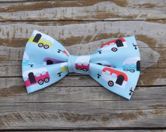 May include: A light blue bow tie with a pattern of colorful camper vans. The bow tie is made of fabric and has a classic bow tie shape.