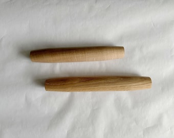 May include: Two wooden rolling pin handles, each with a rounded shape and a natural wood grain. One handle is lighter in color, while the other is a darker shade of brown. The handles are displayed on a white surface.