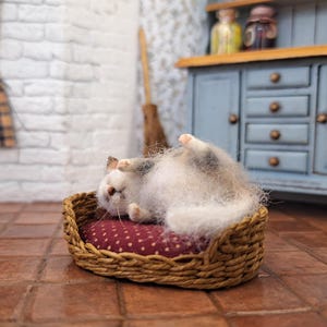 May include: A miniature cat figurine lies in a woven basket on a red polka-dot cushion. The cat has white and grey fur. The scene is set in a dollhouse with a blue cabinet and a brick wall. The basket is made of woven material.