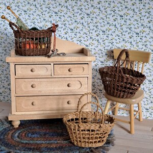 Op de afbeelding: Een miniatuur houten dressoir met vijf laden, een bruine rieten mand gevuld met garen en breinaalden, en twee andere bruine rieten manden. Het dressoir en de manden staan op een houten vloer met een blauw en bruin gevlochten kleed.