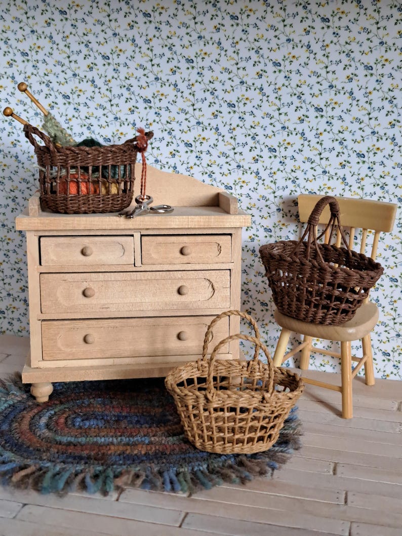 Op de afbeelding: Een miniatuur houten dressoir met vijf laden, een bruine rieten mand gevuld met garen en breinaalden, en twee andere bruine rieten manden. Het dressoir en de manden staan op een houten vloer met een blauw en bruin gevlochten kleed.