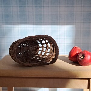 Op de afbeelding: Een bruine geweven mand staat op een houten tafel met drie rode appels ernaast.