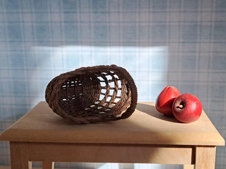 Op de afbeelding: Een bruine geweven mand staat op een houten tafel met drie rode appels ernaast.