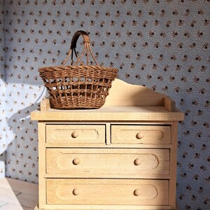Op de afbeelding: Een licht houten dressoir met vier lades en een geweven rieten mand er bovenop. Het dressoir staat op een rood en bruin kleed.