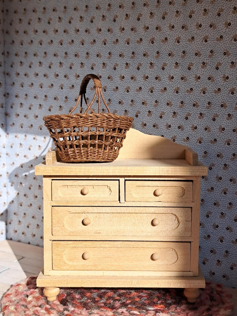 Op de afbeelding: Een licht houten dressoir met vier lades en een geweven rieten mand er bovenop. Het dressoir staat op een rood en bruin kleed.