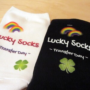 May include: Two pairs of white and black socks with a rainbow and shamrock design. The socks have the text "Lucky Socks - Transfer Day -" printed on them.