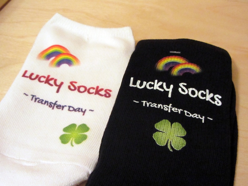 May include: Two pairs of white and black socks with a rainbow and shamrock design. The socks have the text "Lucky Socks - Transfer Day -" printed on them.