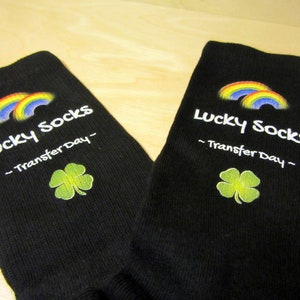 May include: A pair of black socks with a rainbow and shamrock design. The socks have the text "Lucky Socks - Transfer Day -" printed on them.