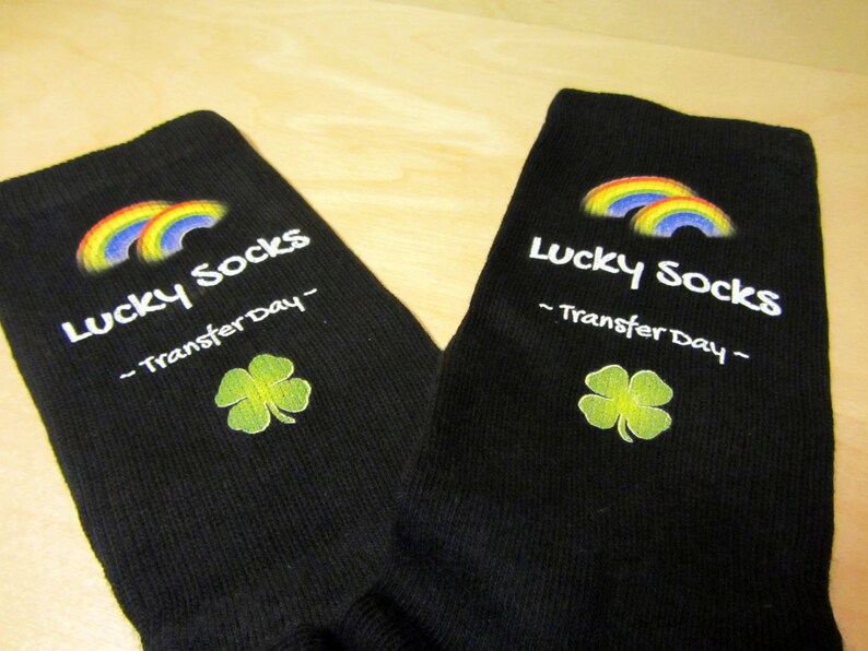 May include: A pair of black socks with a rainbow and shamrock design. The socks have the text "Lucky Socks - Transfer Day -" printed on them.