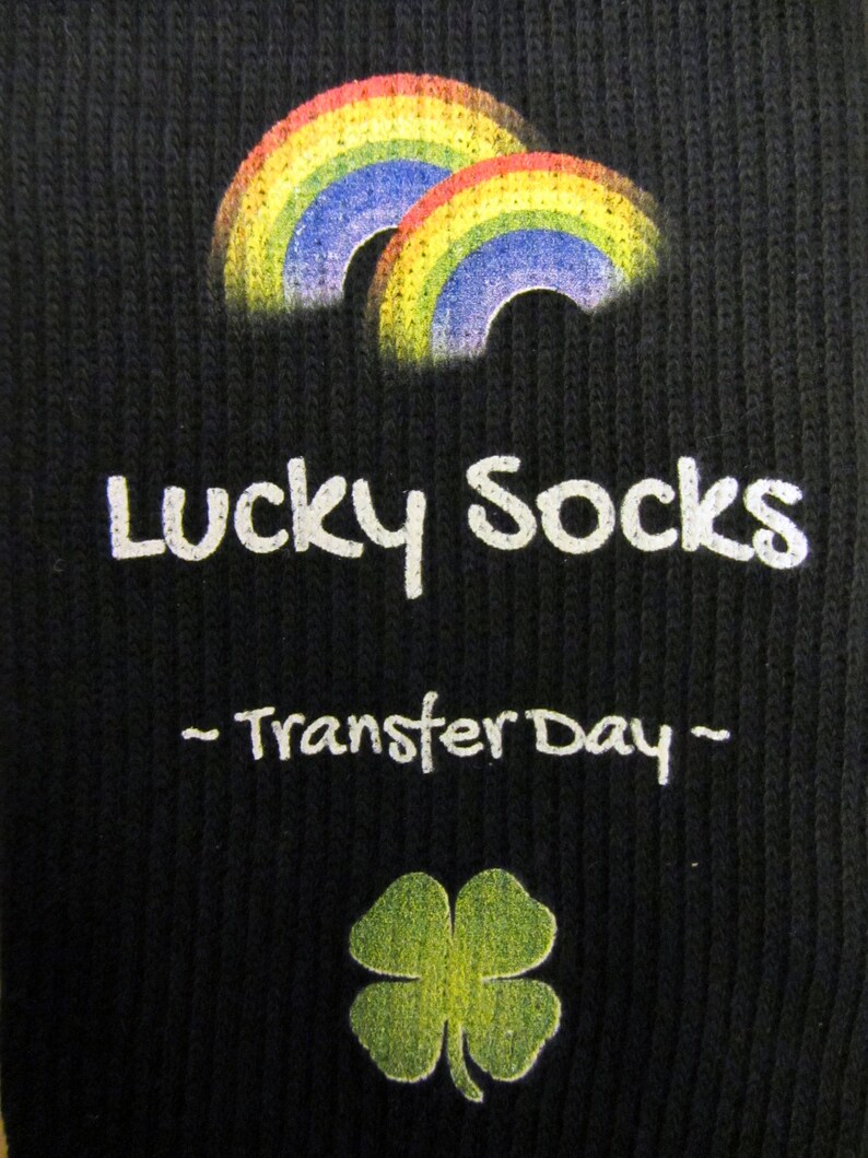 May include: Black socks with a white graphic of two rainbows and the text "Lucky Socks - Transfer Day -" with a green four-leaf clover below.