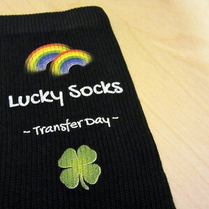 May include: Black sock with a rainbow and a four-leaf clover design. The sock has the text "Lucky Socks - Transfer Day -" printed on it.
