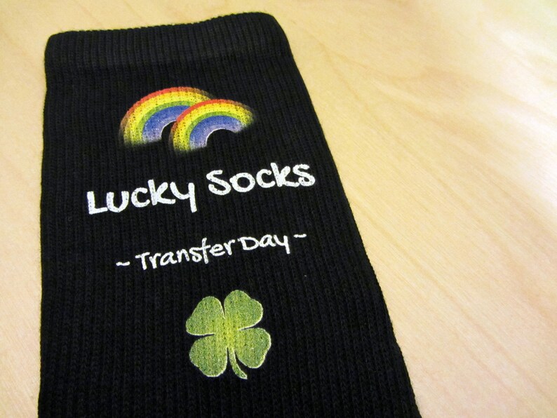 May include: Black sock with a rainbow and a four-leaf clover design. The sock has the text "Lucky Socks - Transfer Day -" printed on it.