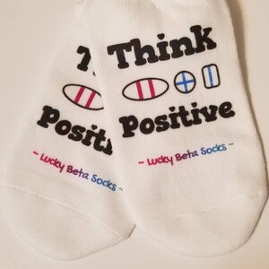 以下が含まれることがあります： 「Think Positive」という文字が書かれた白い足首丈の靴下2足組です。靴下には、虹色の瞳を持つ2つの目のカラフルなグラフィックが描かれています。各靴下の底には「Lucky Beta Socks」という文字が書かれています。