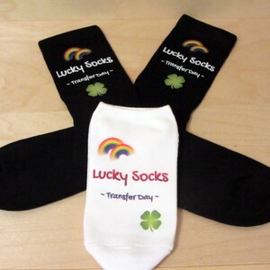 May include: Two pairs of black socks with a white rainbow and shamrock design. The socks have the text "Lucky Socks - Transfer Day -" printed on them.