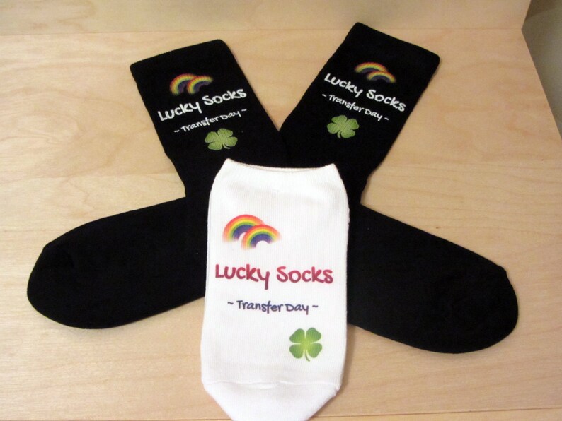 May include: Two pairs of black socks with a white rainbow and shamrock design. The socks have the text "Lucky Socks - Transfer Day -" printed on them.