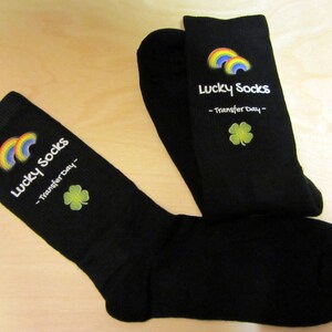 May include: A pair of black socks with a rainbow and shamrock design. The socks have the text "Lucky Socks - Transfer Day -" printed on them.
