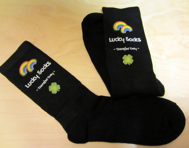 May include: A pair of black socks with a rainbow and shamrock design. The socks have the text "Lucky Socks - Transfer Day -" printed on them.