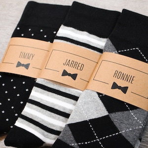 May include: Three pairs of socks with different patterns, each with a brown paper tag with a name and a bow tie graphic. The names on the tags are Timmy, Jarred, and Ronnie.