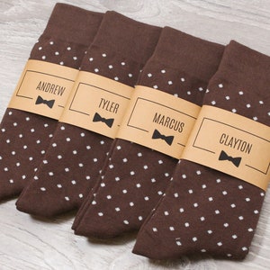 Peut inclure: Quatre paires de chaussettes marrons à pois blancs. Chaque paire est munie d'une étiquette en papier brun avec un nœud papillon blanc et un nom imprimé dessus : Andrew, Tyler, Marcus et Clayton.