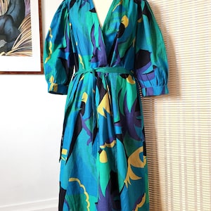 Op de afbeelding: Een vintage jurk met een opvallend abstract patroon in tinten van turquoise, blauw, groen, geel en zwart. De jurk heeft een V-hals, korte mouwen en een bijpassende riem. De jurk wordt op een mannequin gepresenteerd.