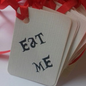 May include: A stack of white gift tags with red ribbon. The tags have the words "Eat Me" printed in black.