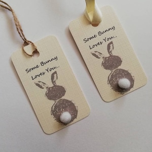Może przedstawiać: Dwie prostokątne etykiety prezentowe z motywem królika i napisem "Some Bunny Loves You...". Etykiety są beżowe z wzorem tekstury, brązową ilustracją królika i białym ogonkiem z pomponu. Jedna etykieta ma sznurek, druga kremową wstążkę.