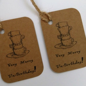 May include: Two brown gift tags with twine attached. Each tag features a black and white illustration of a stack of teacups and a saucer. The text "Very Merry Un-Birthday!" is printed on each tag.