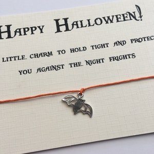 Op de afbeelding: Oranje koordarmband met een zilveren vleermuisbedel. De kaart die aan het armband is bevestigd, luidt "Happy Halloween! Klein charme om vast te houden en je te beschermen tegen de nachtmerries."