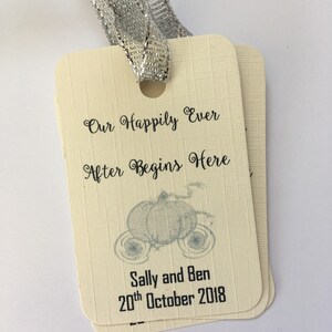 Könnte beinhalten: Elfenbeinfarbene Hochzeitsanhänger mit abgerundeten Ecken, mit dem Text "Our Happily Ever After Begins Here" und einer Kutschenillustration. Die Anhänger sind mit einem silbernen Band gebunden und enthalten die Namen "Sally and Ben" und das Datum "20. Oktober 2018".