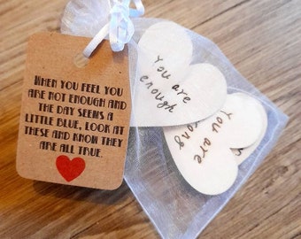 May include: A small gift set featuring a kraft paper tag and wooden hearts. The tag has text and a red heart. The wooden hearts are inside a sheer white bag. The text on the tag reads: "When you feel you are not enough and the day seems a little blue, look at these and know they are all true."