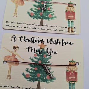 Puede incluir: Dos pulseras de deseos navideños en tarjetas. Cada tarjeta tiene una ilustración de bailarina, un árbol de Navidad y una ilustración de cascanueces. El texto dice "A Christmas Wish from Me to You". Una pulsera tiene un dije de bailarina, la otra un dije de cascanueces.