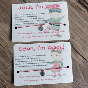 Könnte beinhalten: Zwei rot-weiße Weihnachtskarten mit einer Elfenillustration und einem roten Armband mit einem silbernen Anhänger, auf dem "Glaube" steht. Die Karten enthalten eine Nachricht für Jack und Eden, in der steht, dass der Elf für mehr Spaß und Spiele zurückgekommen ist und Berichte an den Weihnachtsmann über ihr Verhalten senden wird.
