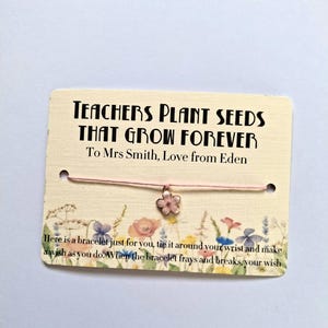 Op de afbeelding: Een roze koordarmband met een bloemvormige bedel is bevestigd aan een kaart. De kaart leest "TEACHERS PLANT SEEDS THAT GROW FOREVER" en "To Mrs Smith, Love from Eden". De kaart is versierd met bloemenillustraties.