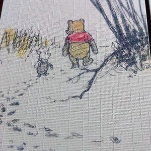 May include: A Winnie the Pooh and Piglet illustration on a white card with a brown background. The illustration shows Winnie the Pooh and Piglet walking away from the viewer. Winnie the Pooh is wearing a red shirt and Piglet is wearing a black shirt. The illustration is surrounded by a brown border.