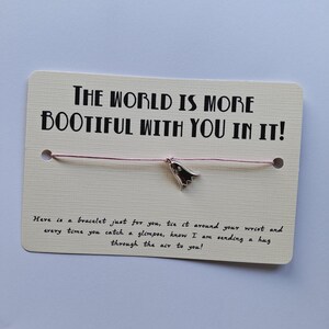 Op de afbeelding: Een roze koordarmband met een zilveren bedel in de vorm van een spook. De kaart die aan de armband is bevestigd, luidt "The world is more bootiful with you in it!" en "Here is a bracelet just for you, tie it around your wrist and every time you catch a glimpse, know I am sending a hug through the air to you!"