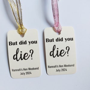 Może przedstawiać: Dwie białe etykiety prezentowe z czarnym tekstem, który brzmi "But did you die?" i "Hannah's Hen Weekend July 2024". Jedna etykieta ma złotą wstążkę, a druga różową wstążkę.