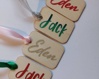 May include: Four wooden gift tags with the names Eden and Jack written on them in different colors. The tags are tied with different colored ribbons.