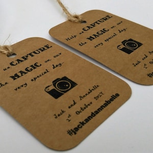 Könnte beinhalten: Zwei braune Papieretiketten mit einem Kamerasymbol und dem Text "Help us CAPTURE the MAGIC on our very special day..." Die Etiketten enthalten auch die Namen "Jack und Annabelle" und das Datum "2. Oktober 2017" sowie den Hashtag "#jackandannabelle".